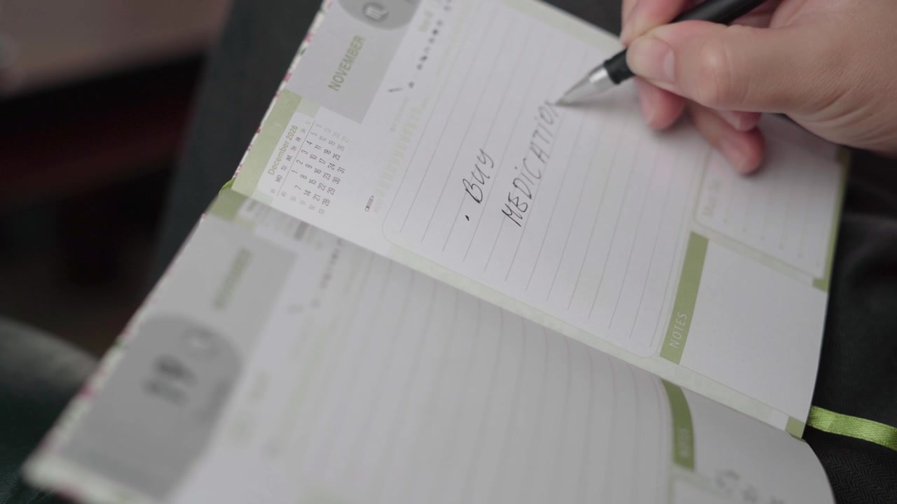 Close-up of a person writing a pharmacy visit reminder to buy medicine in a November paper planner.