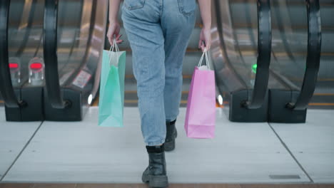 lower body view of lady in jeans and black boots holding two shopping bags stepping onto upward-moving escalator in modern mall setting