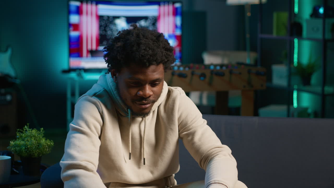 Black remote worker enjoying flexibility and comfort while browsing online on laptop in his stylish apartment decorated with neon lights. Freelance work perfectly captured in a cozy home. Camera A.