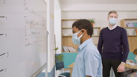 side view of student wearing face mask writing a sentence on blackboard in english classroom while teacher watching him