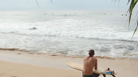 Hombre-Sentado-En-La-Playa-De-Arena-Con-Tabla-De-Surf-Mirando-Las-Olas