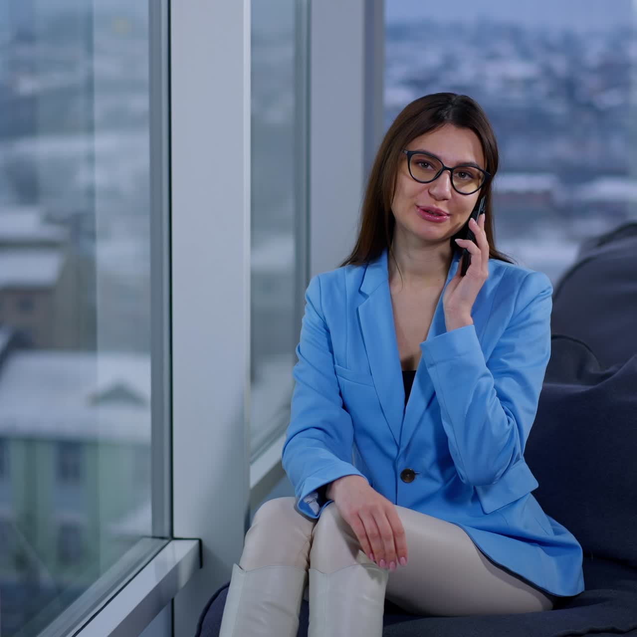 Beautiful young lady in blue jacket sits at the window. Woman speaking on the phone smiling. Blurred backdrop