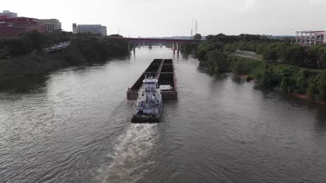 Tugboat-pushes-and-guides-large-commercial-transport-barge-on-river-by-downtown-Nashville-city,-Illinois,-forward-aerial