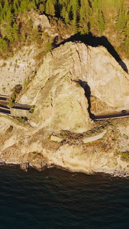 cars running by the highways blocked by the rocks with tunnels inside. rocky wooded shore of lake tahoe, california, usa. aerial perspective. vertical video