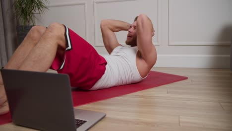 A-confident-blond-man-with-an-athletic-build-and-stubble-in-a-white-T-shirt-and-red-shorts-does-exercises-on-the-mat-to-develop-the-abdominal-muscles-and-watches-a-guide-about-sports-on-his-laptop-in-a-modern-apartment-at-home