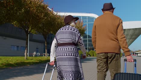 Senior-husband-wife-retirees-tourists-go-to-airport-terminal-for-boarding-with-luggage-on-wheels