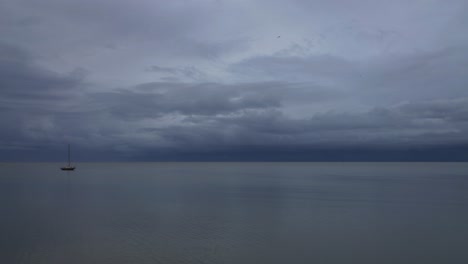 Ein-Einsames-Segelboot,-Das-An-Einem-Sehr-Bewölkten-Tag-Auf-Dem-Ruhigen-Und-Glasigen-Wasser-Schwimmt