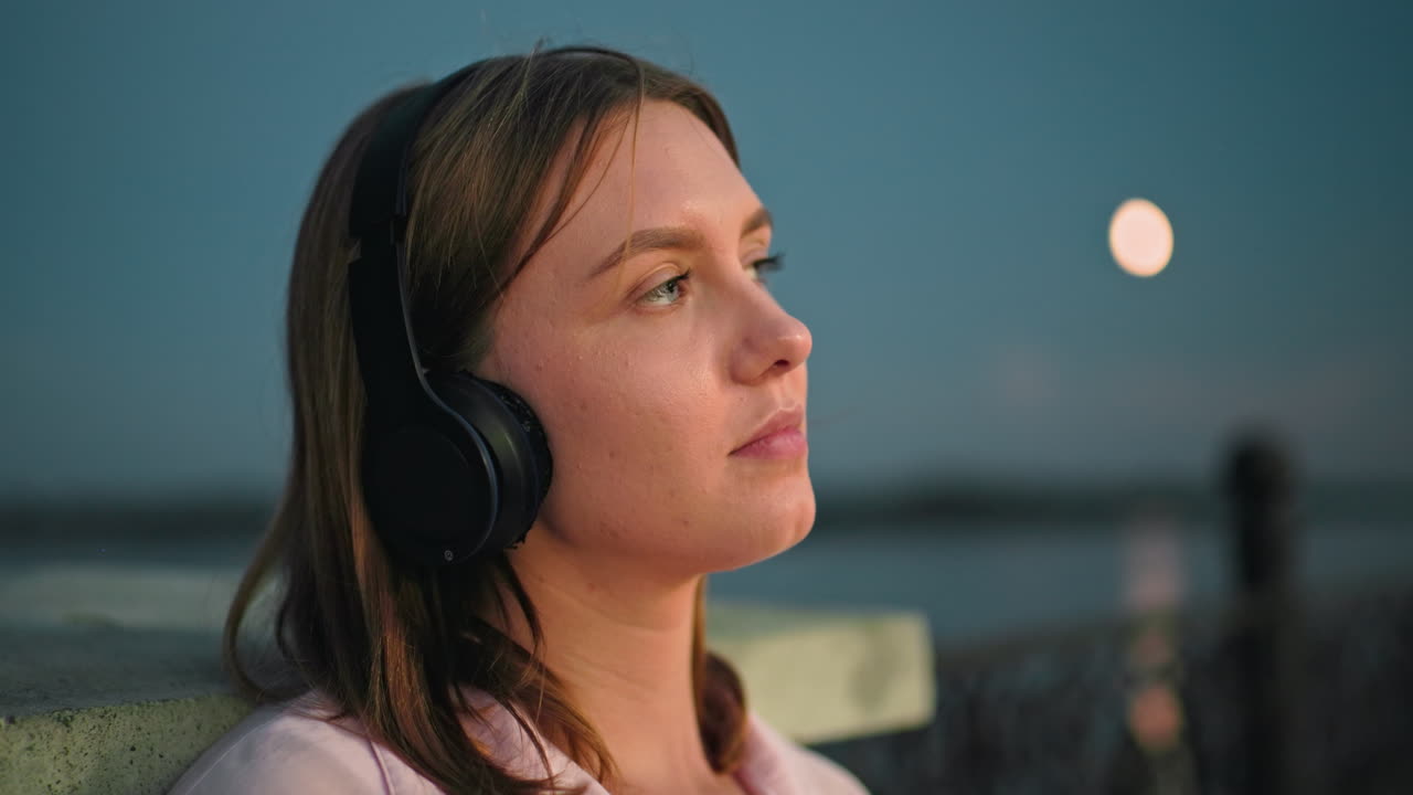 primer plano de una chica con una camiseta rosa y auriculares descansando en la valla mirando hacia arriba pensativo con la luna en el fondo, entorno al aire libre con atmósfera tranquila de la noche