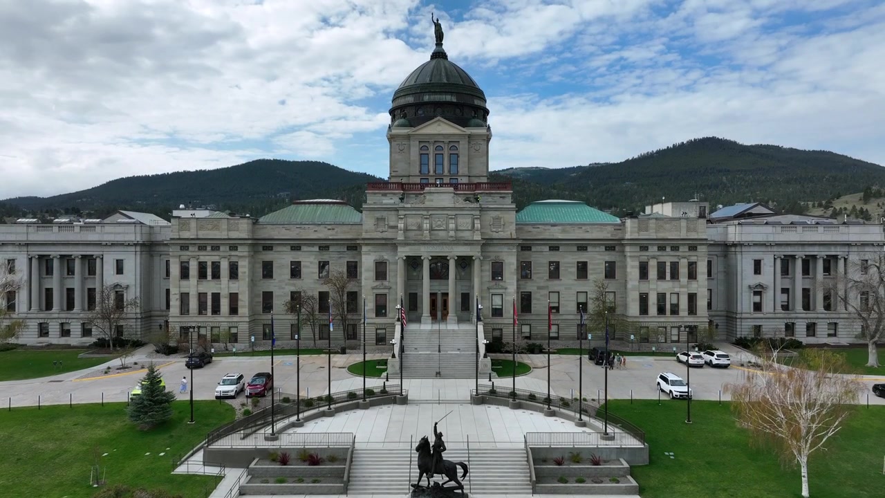 Edifício do capitólio estadual com cúpula ergue-se atrás de monumento equestre de bronze e gramado bem cuidado, ladeado por montanhas arborizadas sob céu parcialmente nublado. Helena, Montana, EUA. Tomada ampla de ascensão de drone aéreo