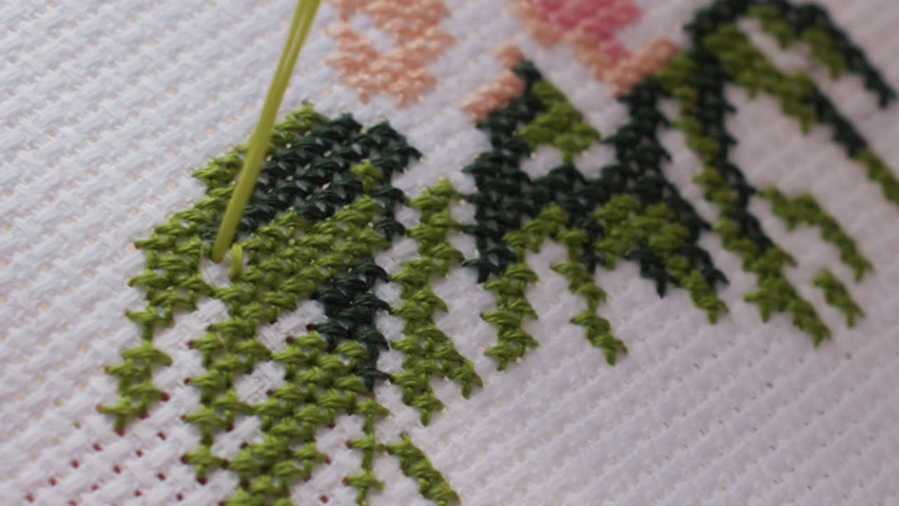 Close up designer embroidering white canvas, needle with green thread passing through cross stitch, leafy pattern forming, fine texture visible, careful craft captured under soft light