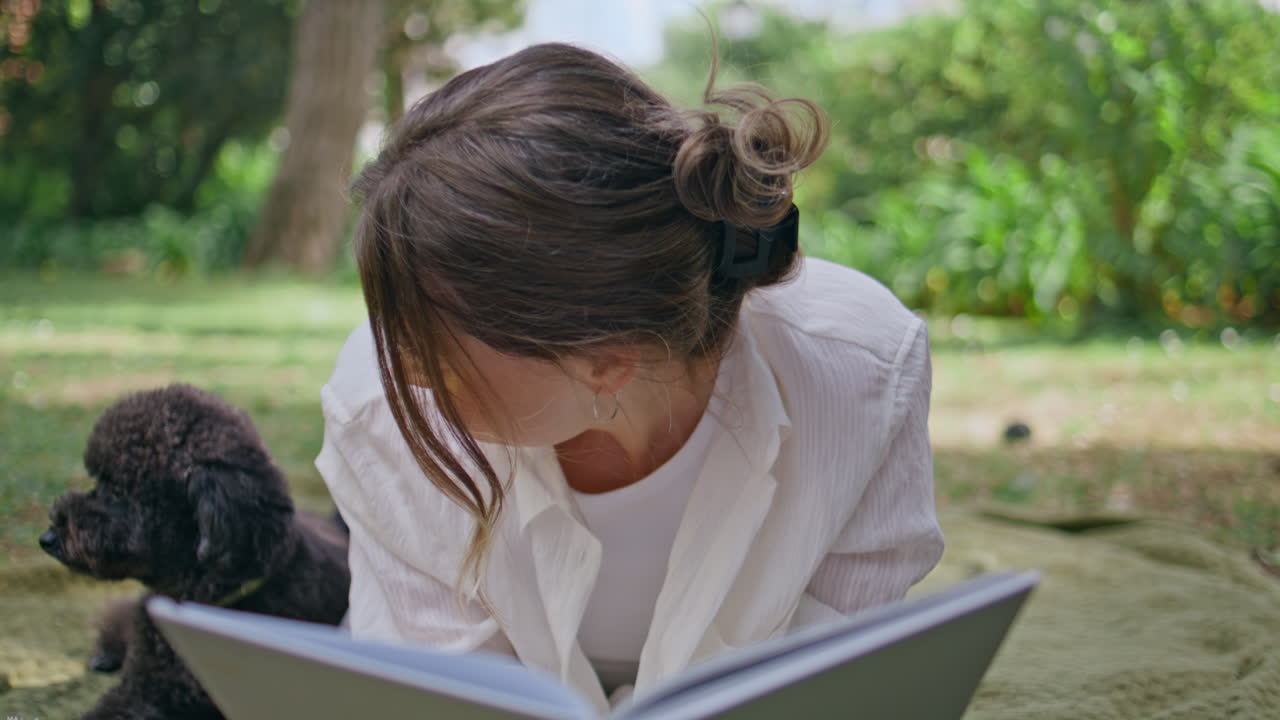 Girl relaxing meadow dog closeup. Happy woman reading book on picnic blanket