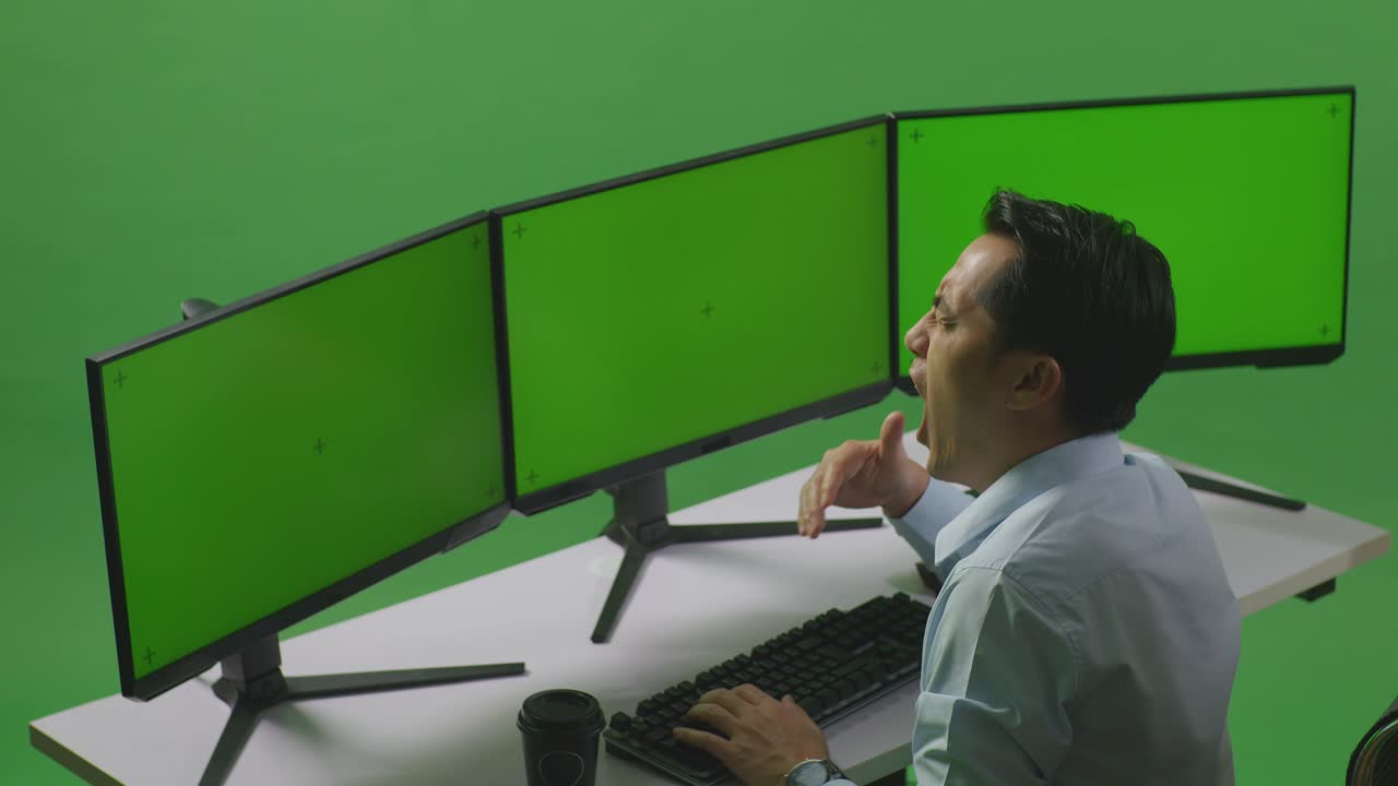 녹색 스크린 스튜디오에서 모의 여러 컴퓨터 모니터와 함께 작업하는 동안 피곤한 아시아 남자의 측면 뷰