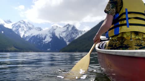 young man with yellow life jacket in calm lake rowing towards snowy mountains