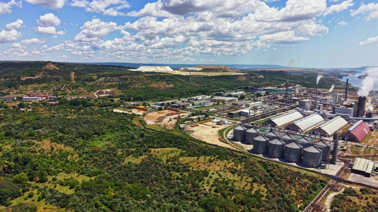 Udsigt til industriområde med lagerstrukturer og røg i baggrunden, beliggende nær grønne bakker og blå himmel.