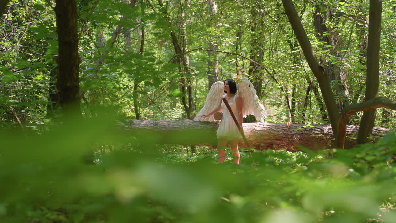 Actress dressed in divine costume with white wings stands near fallen tree in bright green forest, gently resting hand on trunk while surrounded by glowing sunlight