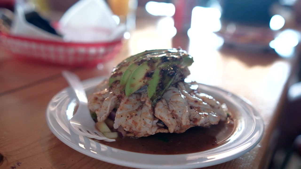 Vista de perto de uma tostada crocante com aguachile de camarão fresco, abacate fatiado, tempero de pimenta e molho, servida em um prato branco dentro de um restaurante mexicano de frutos do mar.