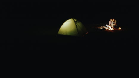 romantic couple relaxing by the campfire at night standing next to a tent in which the light shines