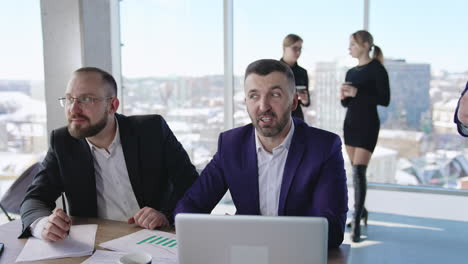 two male businessman talk sitting at desk in front of laptop. they look at something in computer and think it over. close up. ladies chatting and drinking coffee at the backdrop.