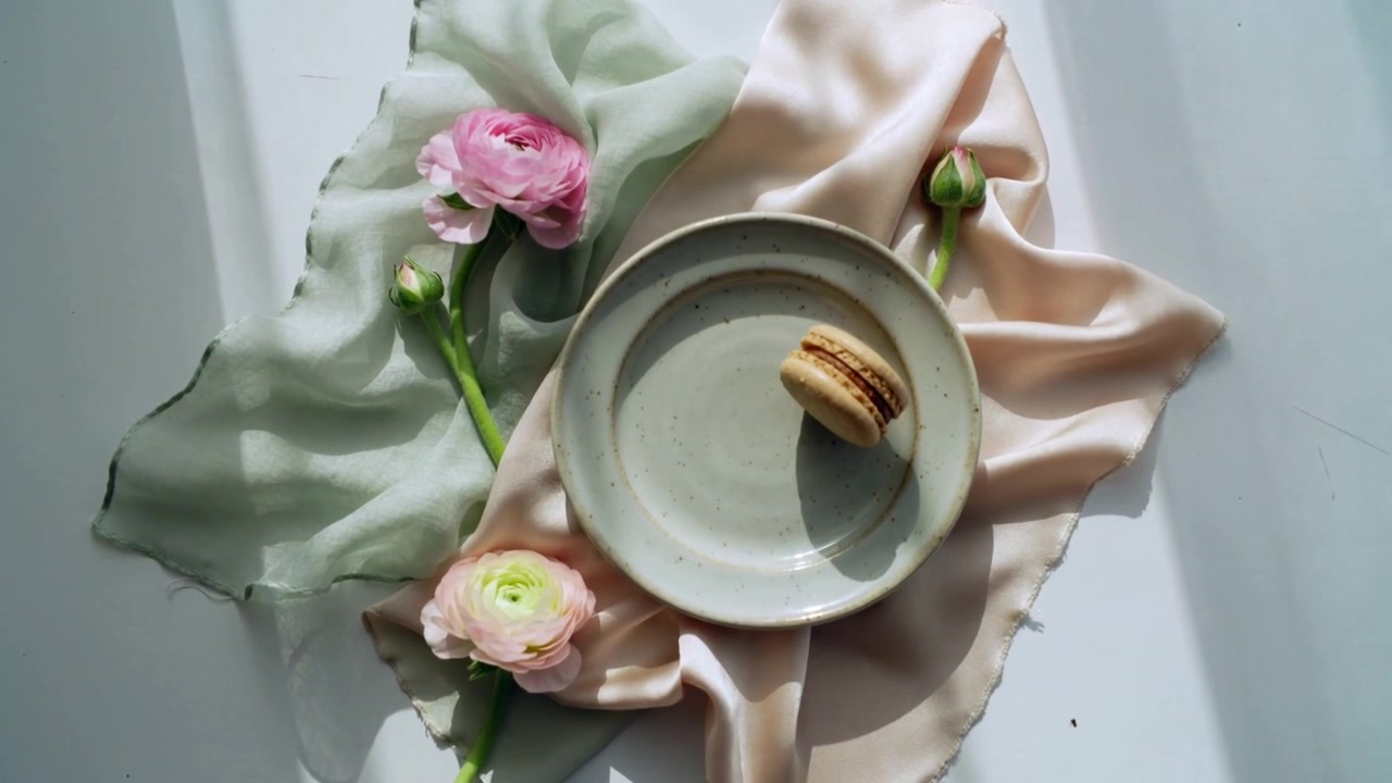 La telecamera si muove stringendo l'inquadratura, evidenziando un macaron su un piatto, ranuncoli rosa e seta.