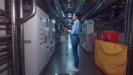 full body side view of asian female professional worker standing with tablet in engine control room, she observes by looking around before she come to concentrating with tablet