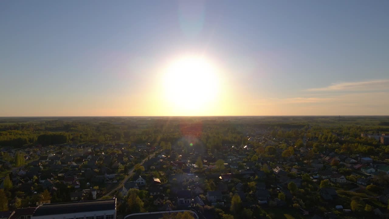 Drone footage capturing a peaceful sunset over a vast landscape featuring a small city and forested areas. The camera slowly descends maintaining symmetry with the sun at the center.