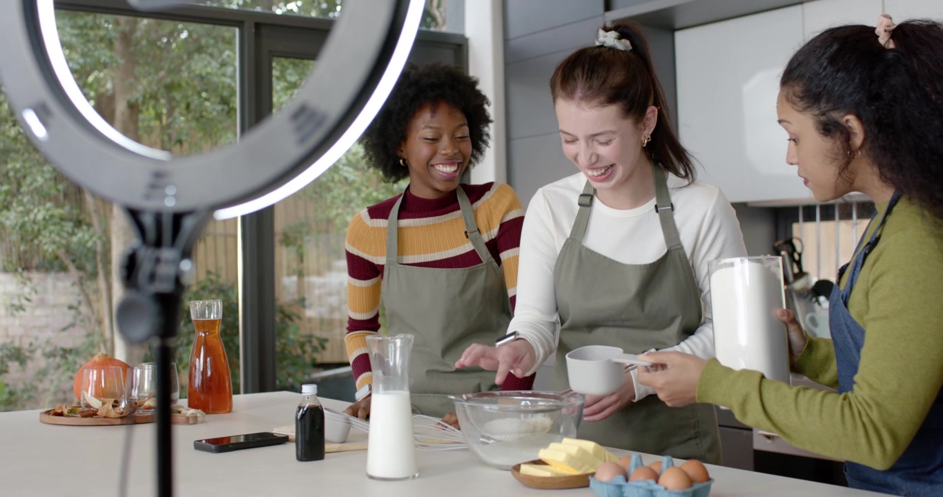 Amiche di diverse etnie che filmano sull'isola della cucina, con una ring light in movimento, mentre versano la farina in una ciotola.