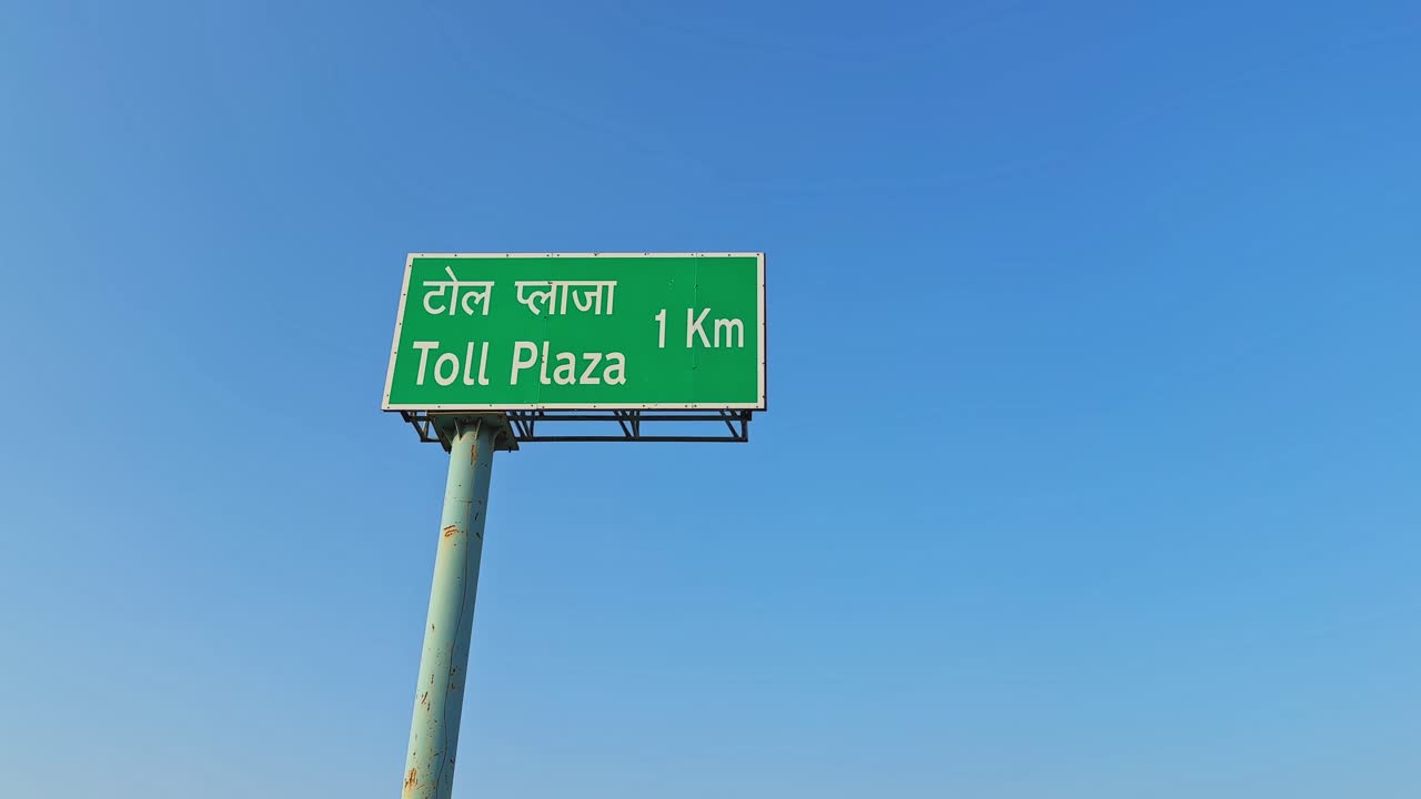 Cartello stradale verde che mostra “Toll Plaza 1 Km” in hindi e inglese, montato su un palo alto contro un cielo azzurro limpido in una composizione statica minimale.