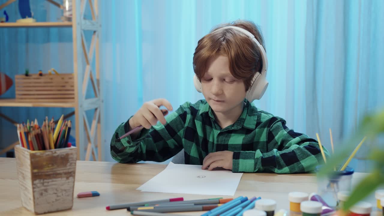 niño de escuela sentado en la mesa en la habitación, dibujando con lápiz en el álbum y escuchando música en grandes auriculares blancos. niño adolescente está involucrado en la creatividad. de cerca. cámara lenta lista, 4k a 59,94 fps