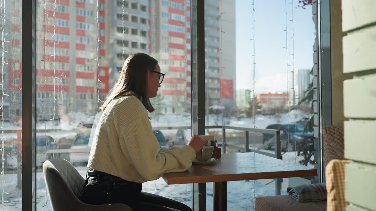 Lonely lady stirs tea slowly while seated by large window in cozy cafe with snowy urban background. Winter light filters in, casting soft shadows