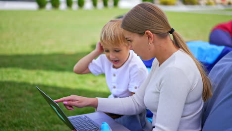 long-haired blonde woman and her little son sitting outdoors. both look at computer discussing something cheerfully. side view.