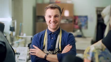 male clothing designer in sewing shop folds his hands and looks at the camera with a smile