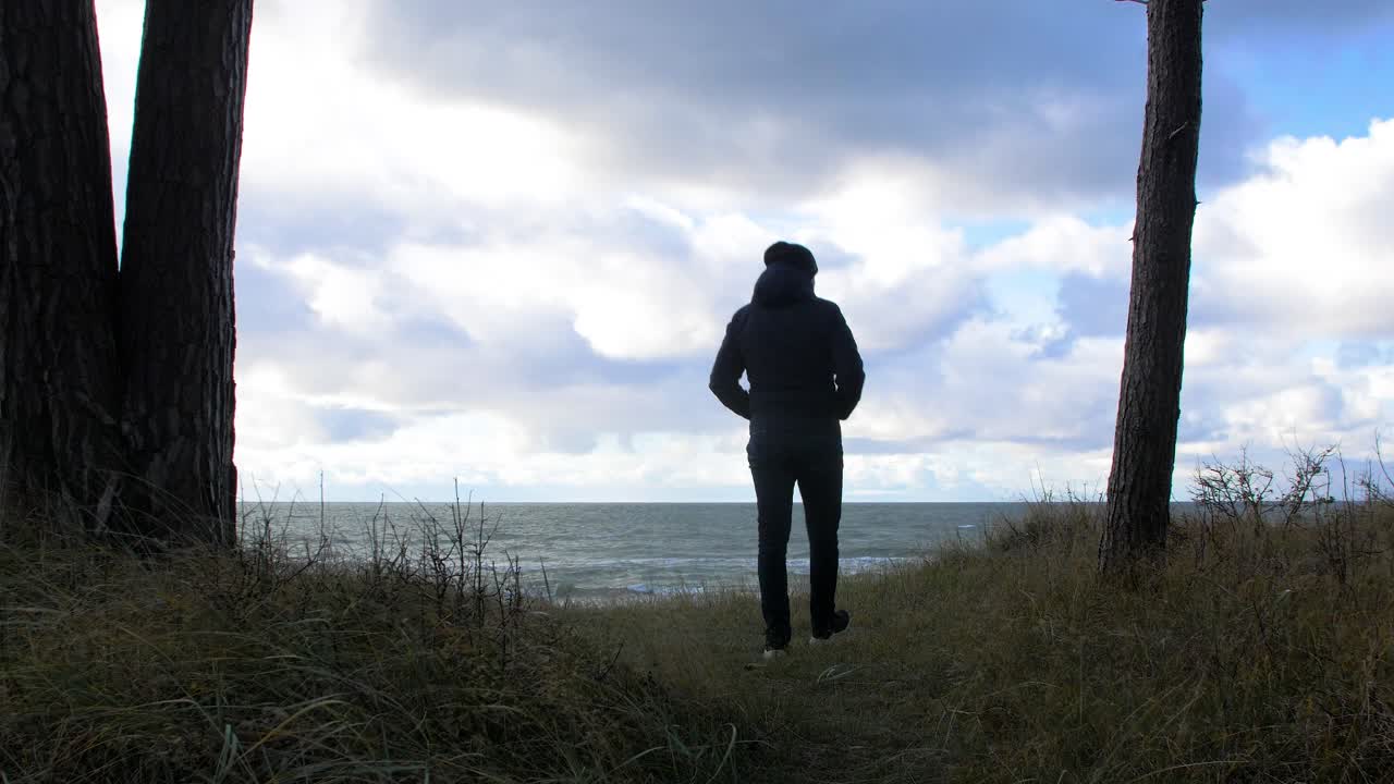 vista trasera de un hombre caucásico explorando el bosque costero nórdico, hombre caminando solo en el bosque de pinos costeros cerca de la playa, día soleado con nubes, concepto de actividad saludable, tiro medio ancho