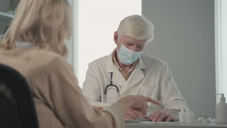 a grey haired, middle aged doctor in a facemask prescribes medicines to a female patient 3