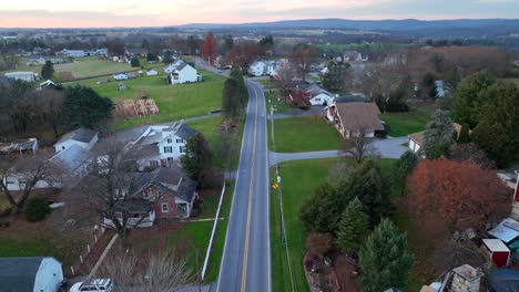 Dusk-over-a-rural-town-with-a-road-flanked-by-houses-and-a-red-barn,-mountains-in-distance