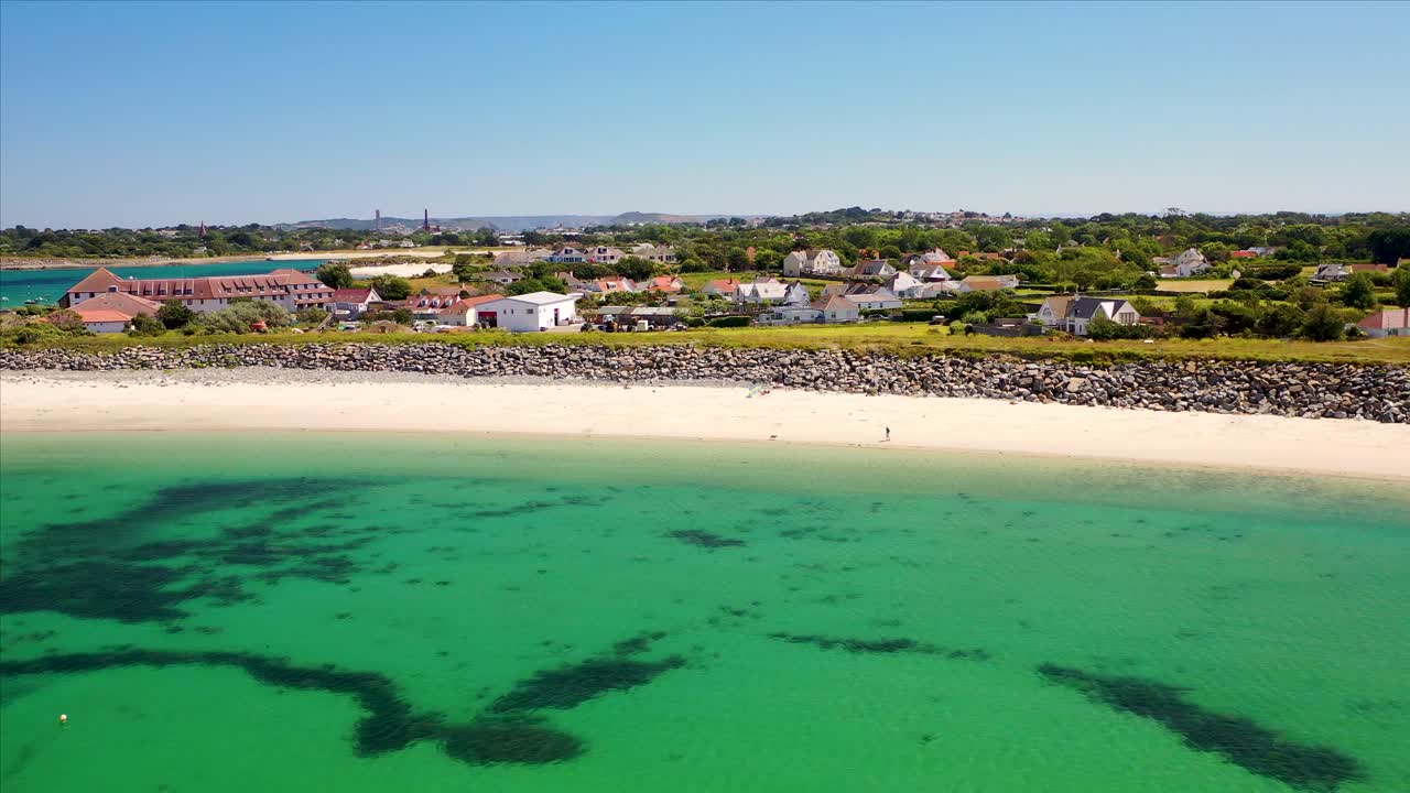 sweeping drone footage of Guernsey coastline, stunning golden beach and clear calm sea on sunny summer day and only one man and his dog on the beach.Cottages and Peninsula Hotel in the background.
