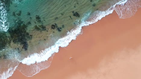 Woman-on-the-pristine-beach-staring-at-beautiful-blue-ocean-water,-birds-eye-view-rising-shot