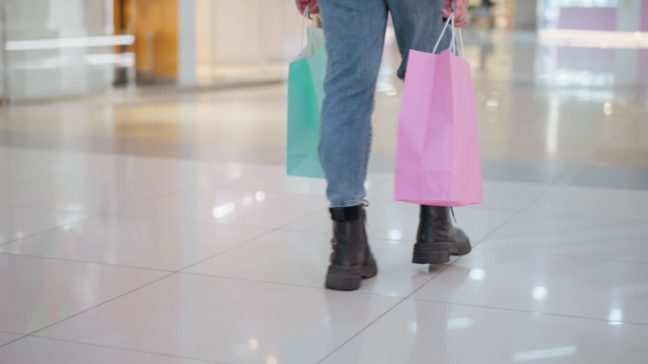 청바지 바지와 검은 부츠를 입은 사람이 쇼핑백을 들고, 반사와 함께 우아한 쇼핑몰 타일에서 고, 다른 쇼핑몰을 지나가며, 바쁜, 활기찬 쇼핑몰 분위기를 보여줍니다.