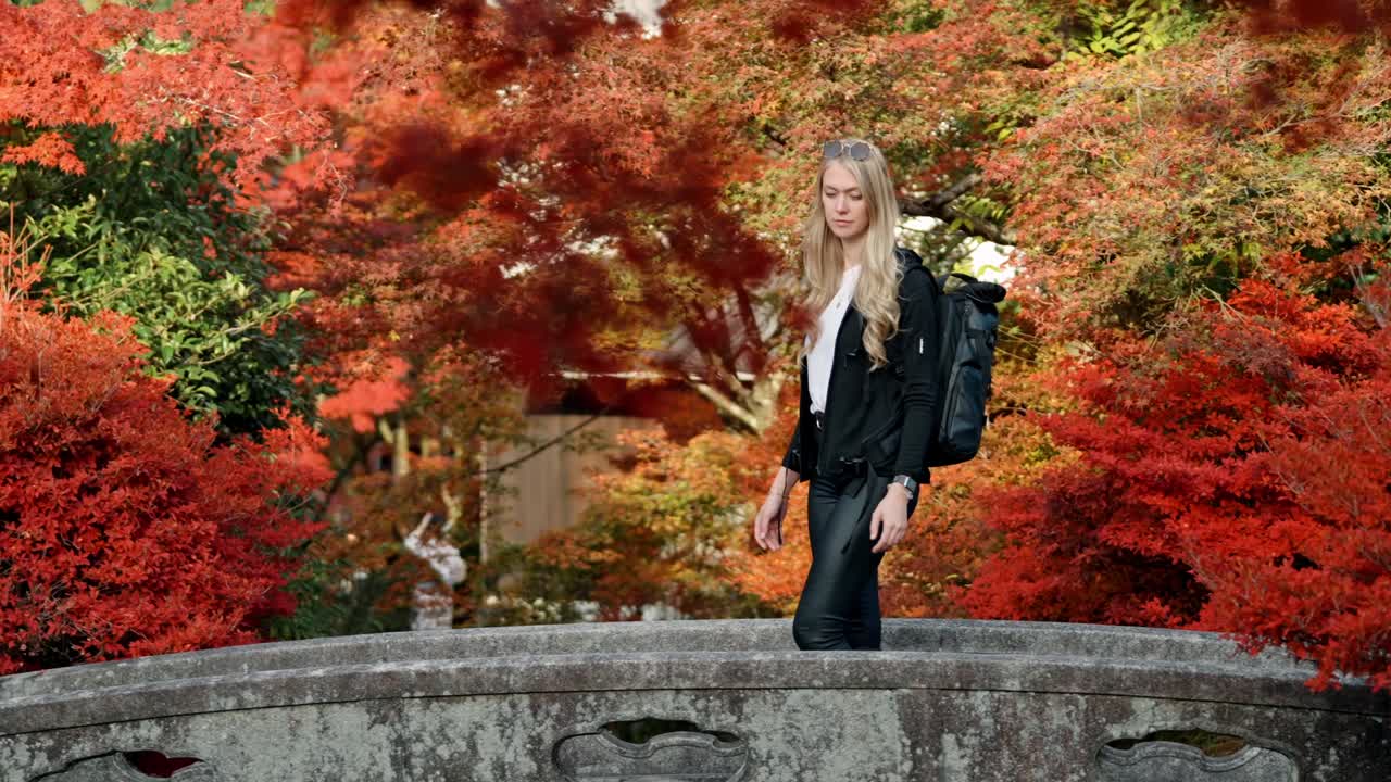 A young woman gracefully walks across a stone bridge at Kiyomizu-dera Temple in Kyoto, Japan, surrounded by vibrant red maple foliage.