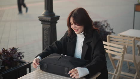 cheerful lady seated outdoors unzips black laptop bag on wooden table, focusing on task, background features blurred outdoor cafe with someone passing by