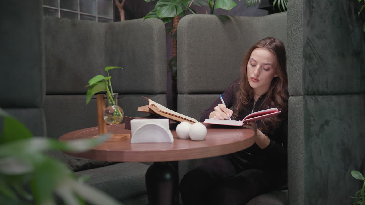 Lady in black outfit and brown shoes writes in pink notebook beside open brown book on round table with white decoration items, seated in green booth surrounded by plants in cozy indoor space