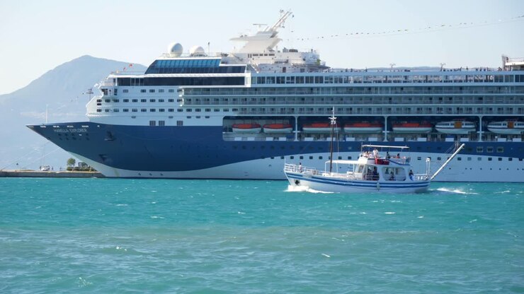 A small boat passes by a large cruise ship, with the mountains of Corfu Island in the background