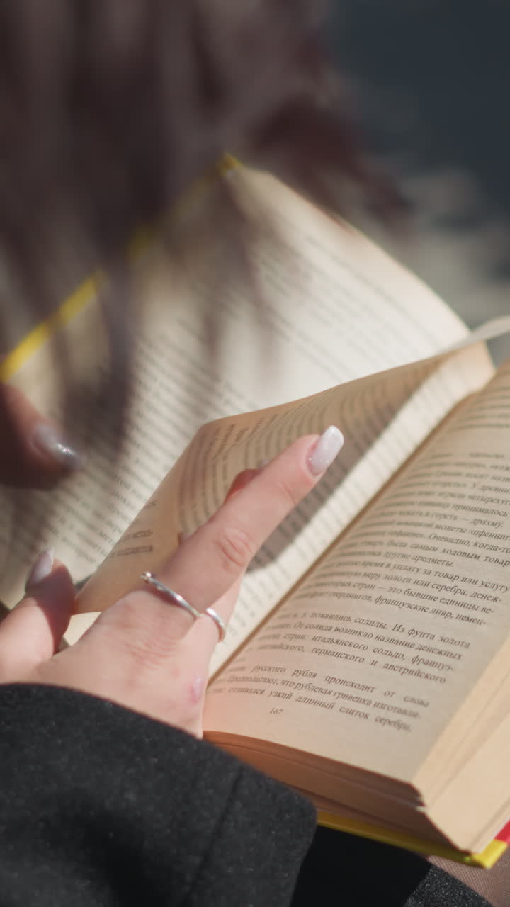 primer plano de una mujer leyendo su libro al aire libre con las manos volviendo la página, sus uñas pintadas y el libro con bordes amarillos está enfocado en el marco, el fondo presenta un entorno urbano y postes de lámparas