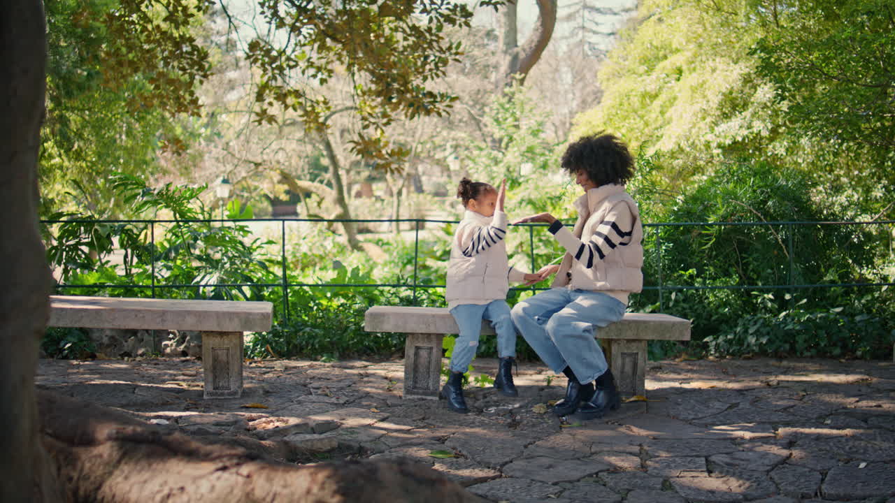 엄마는 손을 박고 딸은 벤치에 앉아 녹색 공원에서 엄마는 소녀를 놀고 있습니다.