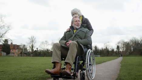 Senior-Woman-Pushing-Senior-Man-In-Wheelchair-Outdoors-In-Fall-Or-Winter-Park