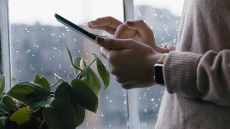 Cerrar-Las-Manos-De-Una-Mujer-Usando-Una-Tableta-Digital-Navegando-Mensajes-En-Línea-Leyendo-Las-Redes-Sociales-Disfrutando-De-Un-Dispositivo-Móvil-Con-Pantalla-Táctil-Parado-Junto-A-La-Ventana-Relajándose-En-Casa-En-Un-Día-Frío-Y-Lluvioso