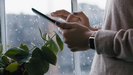 cerca de las manos de la mujer usando una tableta digital navegando por mensajes en línea leyendo redes sociales disfrutando de un dispositivo móvil con pantalla táctil de pie junto a la ventana relajándose en casa en un día frío y lluvioso