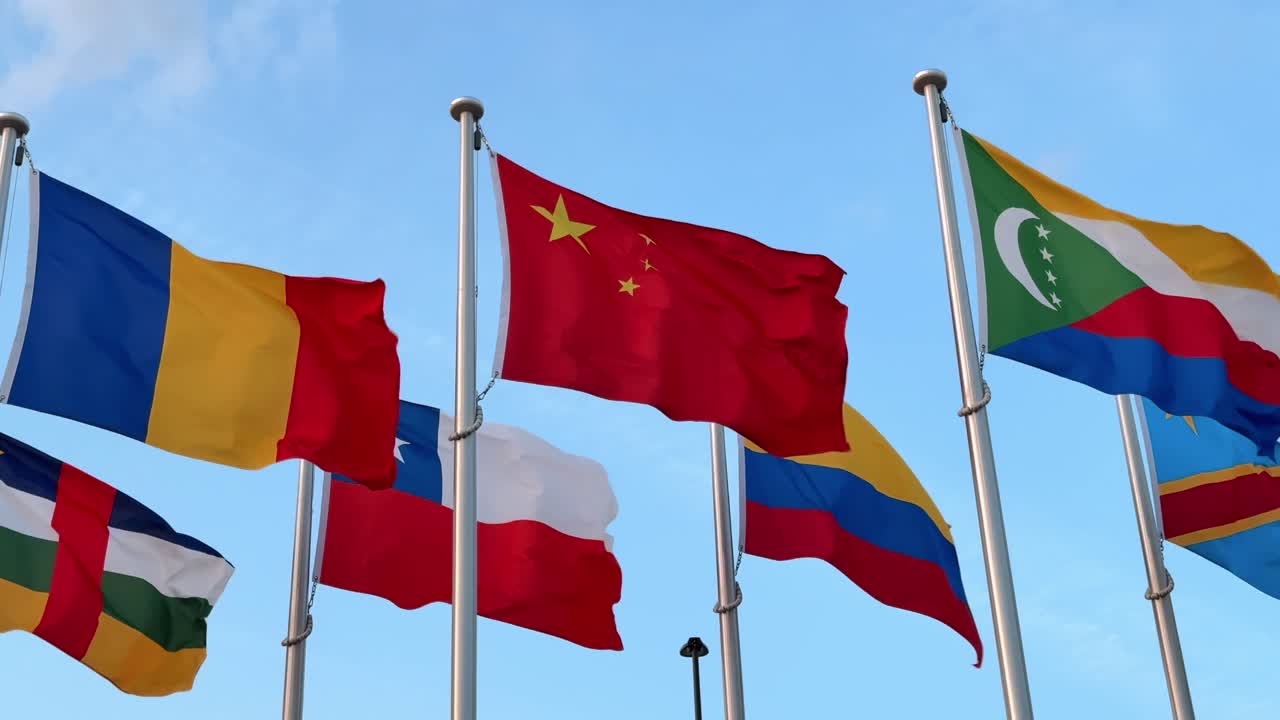 A low-angle shot of vibrant national flags waving in the clear blue sky, with the Chinese flag prominently in the center, symbolizing international presence