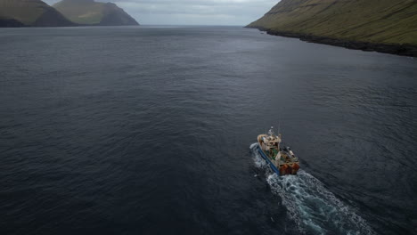 aerial view at close range and with tracking from the back of a fishing boat that sails through a fjord in the faroe islands and where the great mountains can be seen