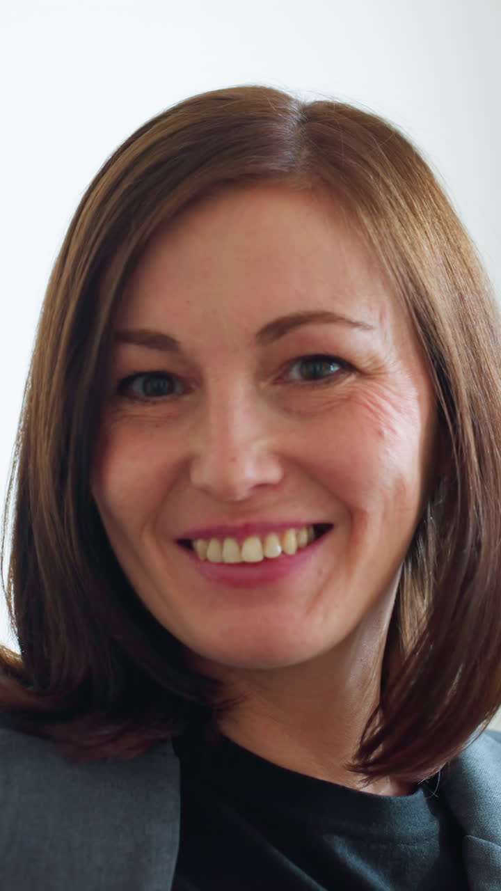 Professional woman in gray blazer sitting comfortably on couch in bright office space with indoor plants, smiling softly and exuding calm confidence in relaxed business environment with natural light
