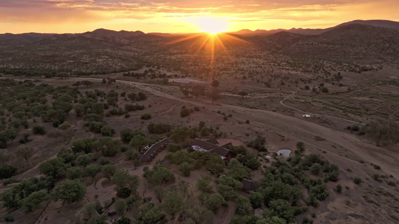 Filmagem de drone de um nascer do sol dourado sobre uma propriedade rural namibiana, luz quente se espalhando por estradas de terra, árvores esparsas e deserto de planalto